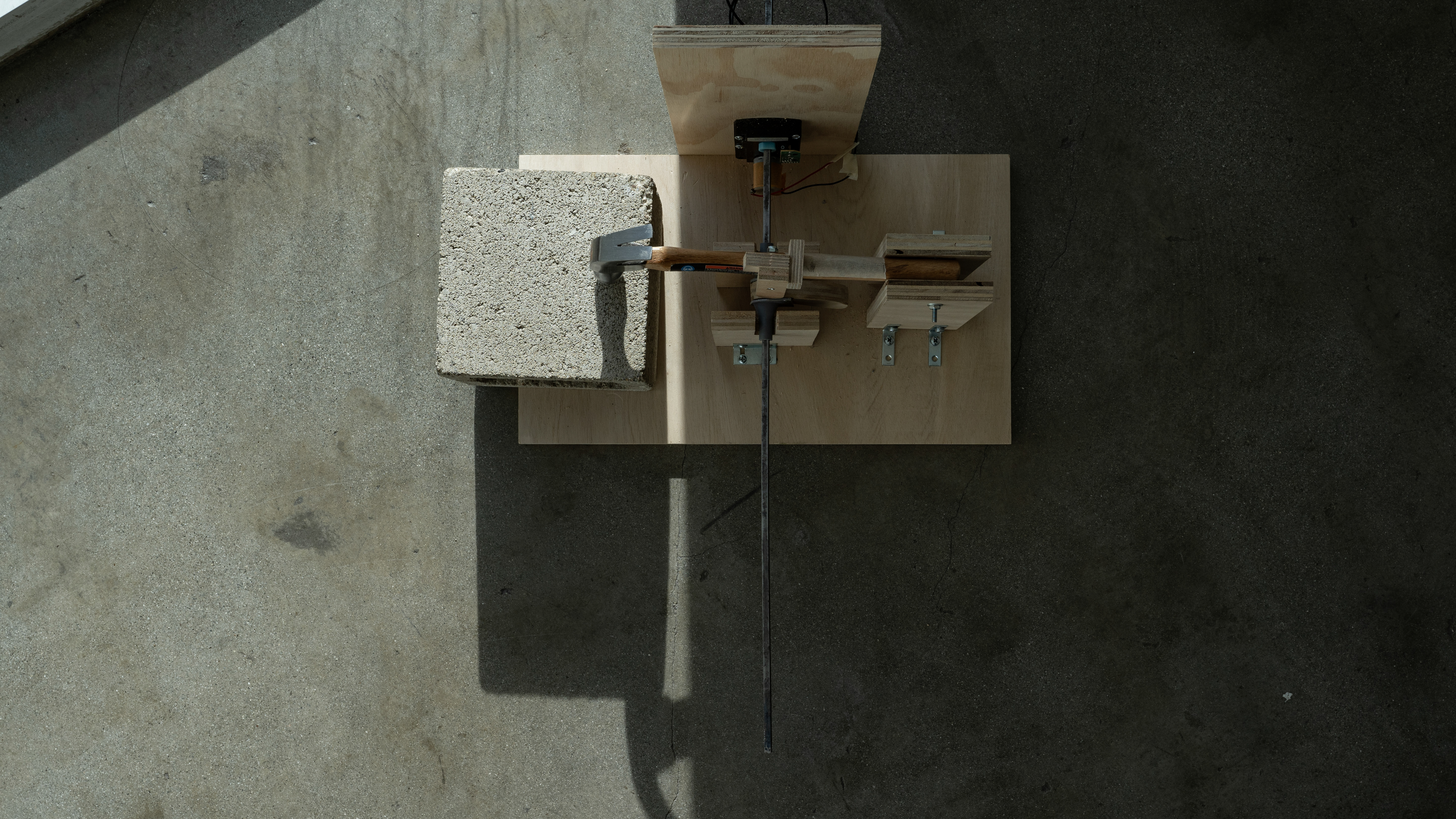 A sculpture consisting of a hammer assembeled by wood with electronics, where the hammer strikes a cinderblock, in dramatic lighting.